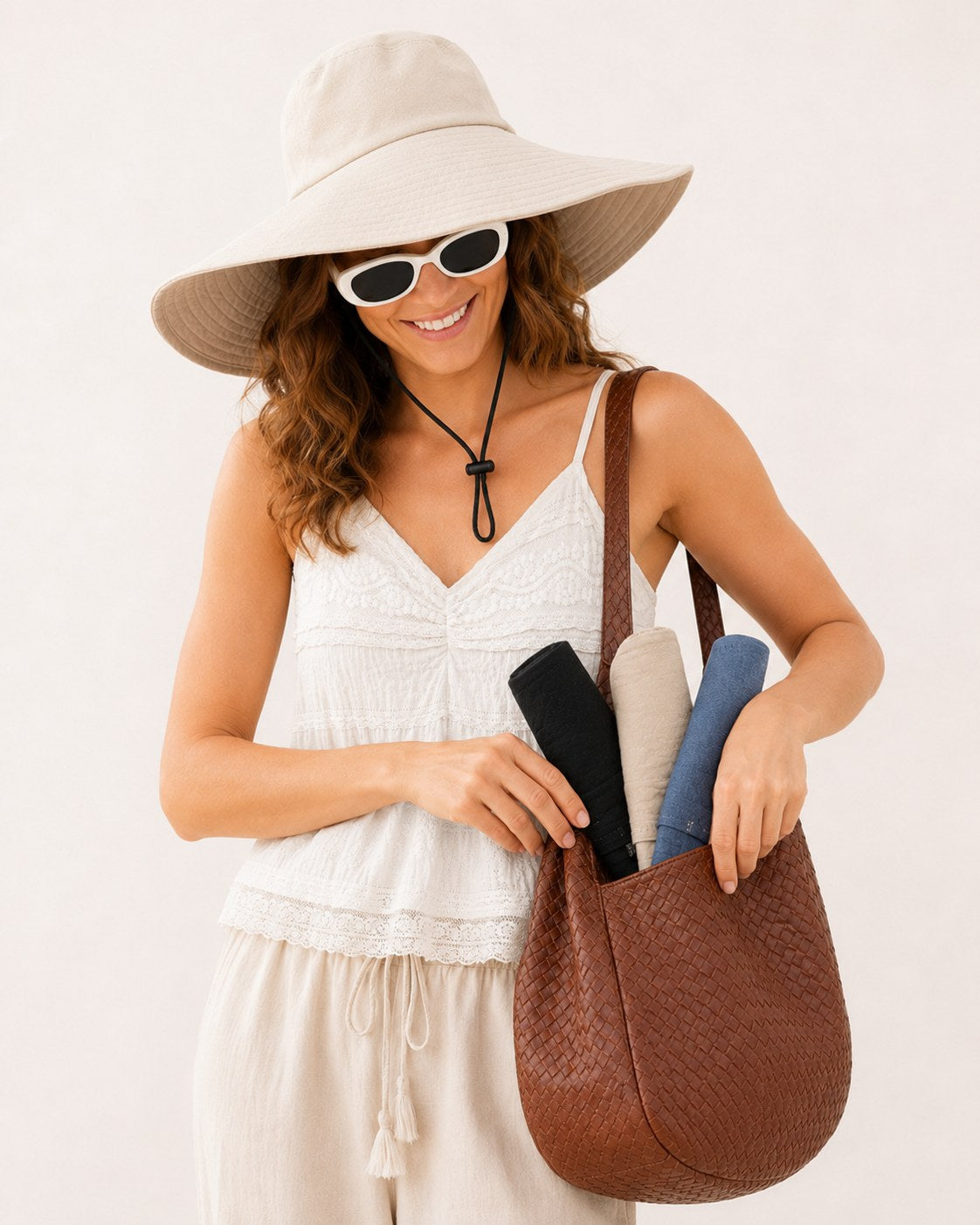 Woman in beige wide-brim sun hat with sunglasses, packing rolled hats into a tote bag