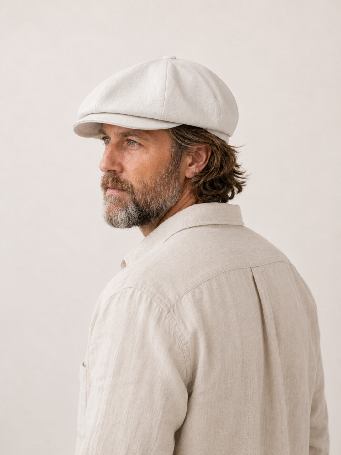 A man in profile wearing a light beige linen flat cap and a matching light beige linen shirt, photographed against a neutral off-white background.