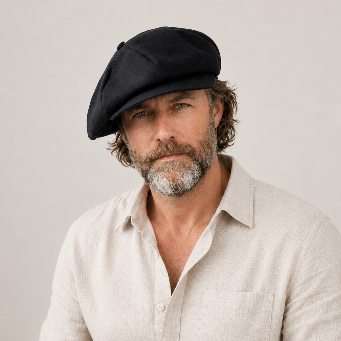 A man wearing a black newsboy cap and a light beige linen button-down shirt, photographed against a neutral light gray background.