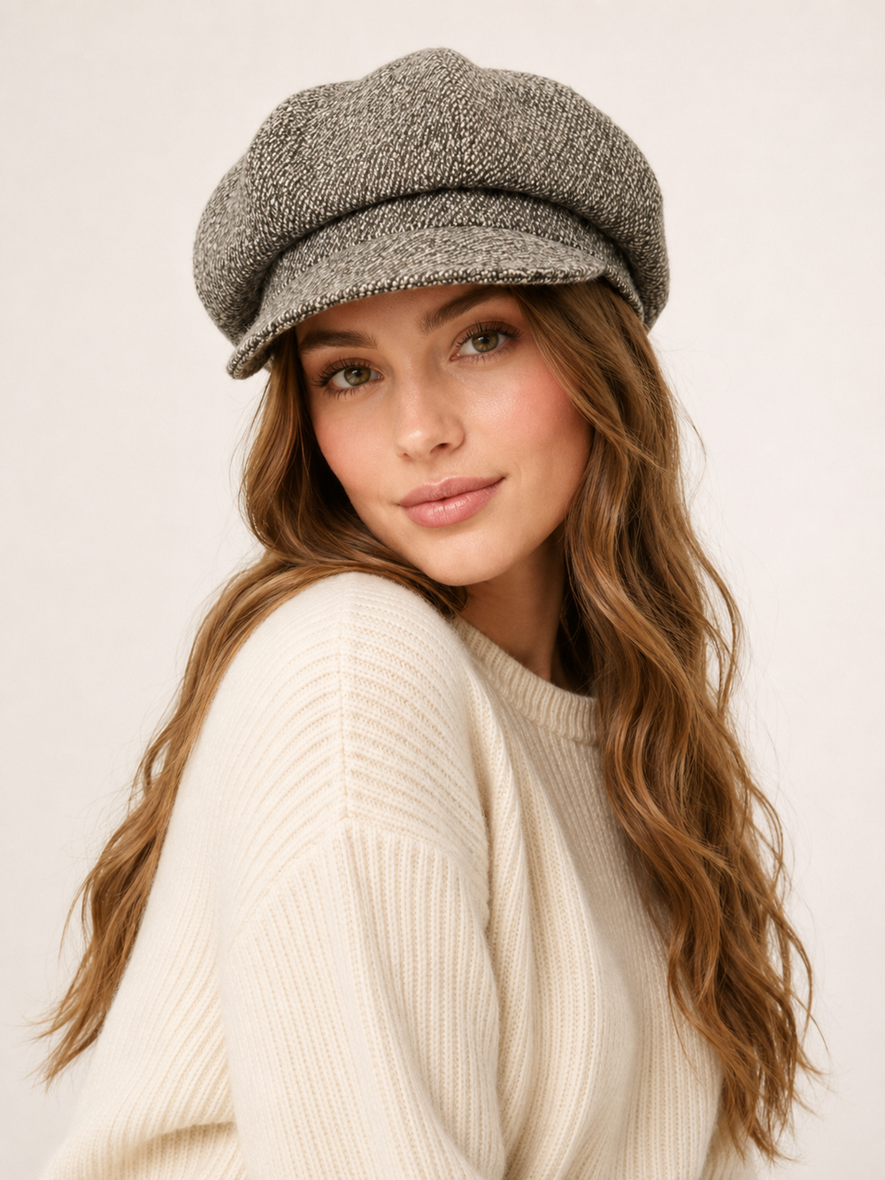A woman wearing a textured gray newsboy cap and a cream ribbed knit sweater, photographed against a plain light background.