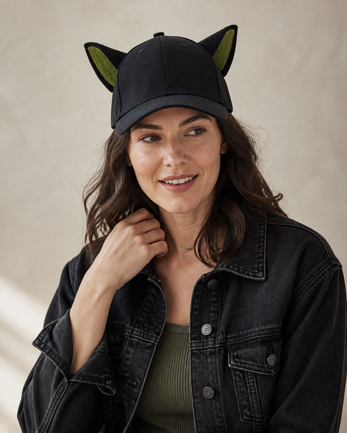 Adult woman wearing the black animal-ear baseball cap with olive-green inner ears in a warm off-white studio