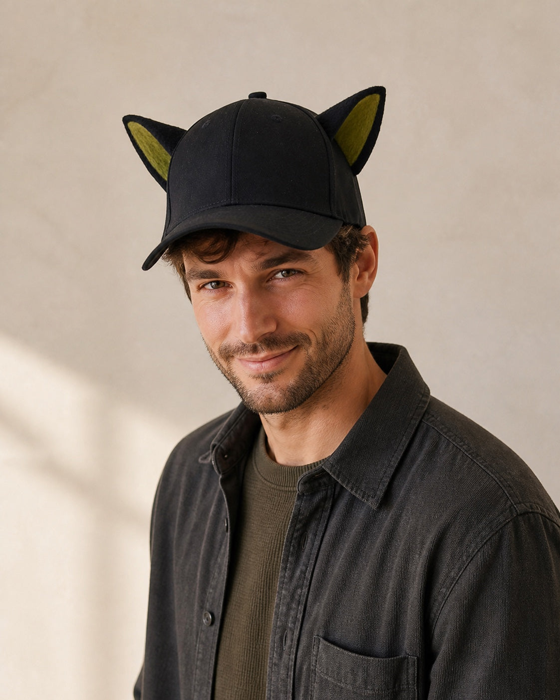 Adult man wearing the black animal-ear baseball cap with olive-green inner ears in a warm off-white studio