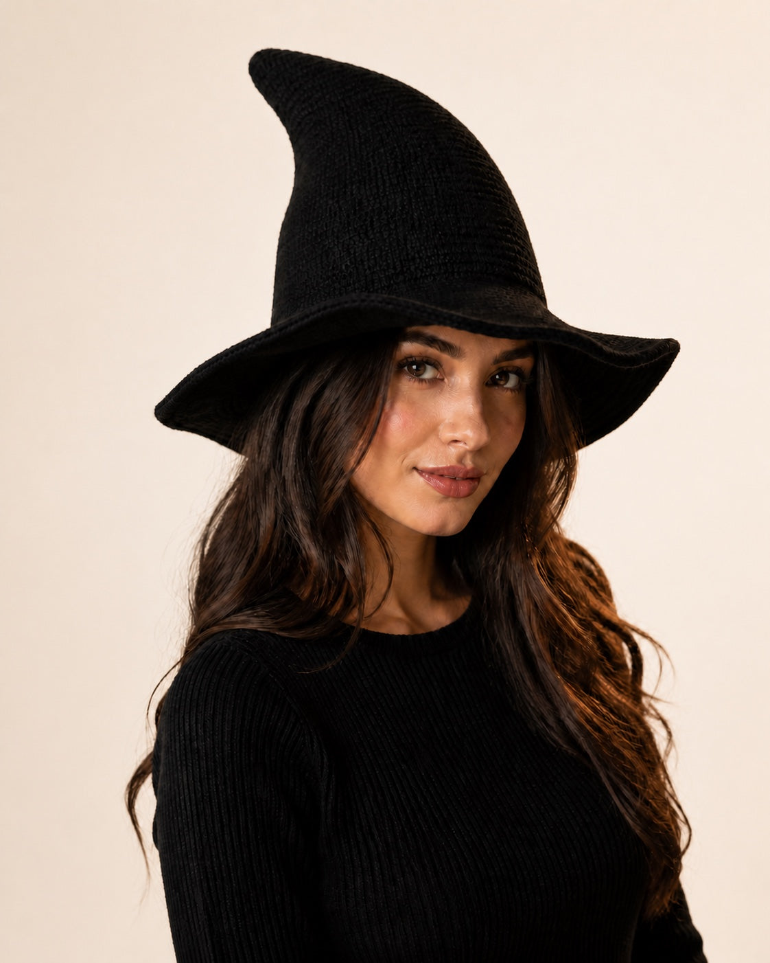 Model wearing a black knitted witch hat against an off-white studio background